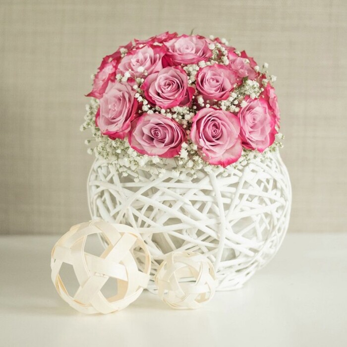 A carefully styled indoor scene features a lush, rounded bouquet of pink roses and white baby's breath poised in a white woven sphere vase, reminiscent of an elegant arrangement you might find in a Clerkenwell design office near Farringdon station. The bouquet forms a near-perfect hemisphere of blooms, with roughly fifteen roses nestled closely together so that their petals overlap and create a rich, layered effect. Each rose displays a soft transition of colour from a subtle, almost ivory blush at the core to a deeper, vibrant pink along the outer edges of the petals, lending a sophisticated two-toned look. Threaded between these roses are fine sprigs of gypsophila, their countless tiny white blossoms adding a light, airy texture that suggests clouds or lace. The flowers are set into a distinctive, spherical container crafted from interlaced white fibres that resemble painted wicker or rattan; its open, criss-cross pattern gives a sense of lightness, as if the bouquet is floating above the white tabletop. In front of and slightly to the left of the vase, two natural-toned decorative spheres rest on the smooth surface. These are made from slim, interwoven wooden strips in a soft beige hue, echoing the woven motif of the vase while introducing a warm, organic contrast to the crisp white surroundings. Behind the arrangement, a softly textured wall in warm beige or off-white creates a gentle, neutral backdrop, similar to the calm interiors of creative studios around Clerkenwell Green. The lighting is bright but diffused, with no harsh shadows, allowing the pink and white tones to appear clean and fresh. The overall impression is of quiet elegance and contemporary charm, ideal for a coffee table, reception desk, or intimate dining setting where subtle romance and thoughtful craftsmanship are appreciated.
