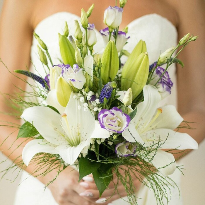 Captured up close in the heart of Clerkenwell, this vibrant bridal bouquet rests gently in a bride's elegantly manicured hands, poised above the smooth sweep of a white, strapless dress. Featuring sweeping white Oriental lilies-some fully unfurled and dusted with golden-yellow stamens, others shyly unopened-this bouquet is a testimony to subtle grandeur. Interwoven are white lisianthus flowers, their soft petals lightly ruffled and edged in cool lavender, alongside a few tight buds peeking through the foliage. Pops of deep, spiky purple florets, reminiscent of local veronica in bloom, provide drama, while clusters of airy white baby's breath add a dreamy texture and balance. Fern-like greenery and bold, dark green lily leaves offer lush structure, with tendrils tumbling naturally to create gentle movement suggestive of Clerkenwell's nearby green spaces. The bride's hands-nails aglow with pale polish-enclose the arrangement with serenity and poise. In the soft, natural lighting that bathes the bouquet, the scene evokes celebration and tranquil anticipation, reflecting both the timeless elegance and creative energy of weddings held in London's historic Clerkenwell.