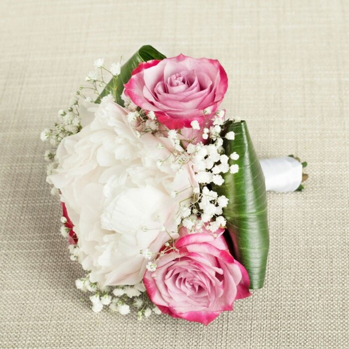 From a slightly elevated angle, this elegant bridal or bridesmaid bouquet evokes the artisanal soul of Clerkenwell, presented against a neutral-textured background akin to the classic fabrics found in local ateliers. The arrangement is woven around a pair of striking pink roses, their color gracefully shifting from vibrant fuchsia and magenta outer petals to soft blush and pale lavender centers, a subtle echo of Clerkenwell's ever-evolving palette. Between them, an opulent white peony takes center stage-its profusion of delicately ruffled petals suggests both abundance and finesse, imbuing the bouquet with a luminous, cloud-like beauty. Sprigs of white baby's breath fill the spaces, imbuing the composition with airy lightness and a romantic softness reminiscent of sunlit strolls through Spa Fields. Carefully folded deep green leaves border the base, offering architectural structure and contrast, as a pristine satin ribbon wraps the stems into a poised handle ready for a walk down a Hatton Garden aisle. Gentle, even lighting enhances the arrangement's depth and the subtle interplay of textures, making it an ideal choice for refined city weddings or celebrations hosted in Clerkenwell's historic venues. This composition is an homage to the neighbourhood's artisanal heritage-timeless, sophisticated, and unforgettable.