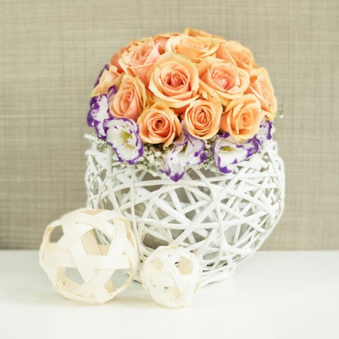 A romantic floral centerpiece sits poised on a smooth white surface, the kind you might find in a light-filled loft overlooking Clerkenwell Green. The arrangement is built around a tightly composed dome of peach and apricot roses, each flower at its peak, with layered petals unfurling in soft spirals. Together they form a rounded, cushion-like globe of colour at the top of the design, radiating warmth and intimacy, as if ready to grace a wedding table or anniversary dinner in EC1. Encircling the base of this rose cluster is a delicate wreath of white lisianthus, whose ruffled petals are finely edged with rich violet and purple tones. These contrasting borders bring depth and gentle drama to the piece, while slender sprigs of white gypsophila are tucked between them, adding a misty, cloud-soft texture that breaks up the dense blooms. The flowers are gathered into a white, broadly spherical woven container, made from smooth wicker or rattan strands that crisscross to form an open lattice. This basket-like vase leans into natural materials yet feels contemporary, echoing the artisan workshops scattered around Clerkenwell's side streets. Placed on the same white surface in front of the arrangement are two decorative orbs crafted from pale, natural wood-toned strips, one larger and the other smaller. Their airy, open weave mirrors the pattern of the vase and introduces a sculptural, minimalist charm. Behind everything, a soft beige fabric backdrop, subtly textured like fine linen, sets a warm, neutral stage. The overall impression is of carefully curated beauty-perfect for modern wedding styling, intimate restaurant tables, or refined home decor in one of Clerkenwell's converted warehouses.