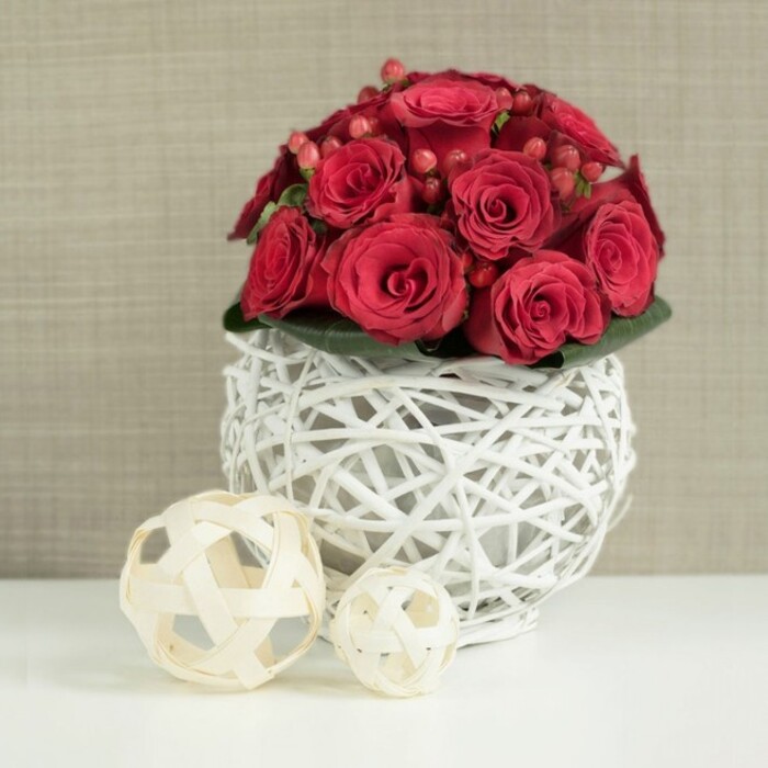 A richly romantic wedding table arrangement of red roses and berries commands the foreground, composed in a perfectly rounded dome that rises confidently from its distinctive vase. The roses themselves are a deep, velvety crimson, each bloom carefully placed so that their petals overlap and interlock, creating a dense, almost seamless carpet of red. Some flowers remain in bud, their outer petals still hugging the centre, while others are more open, revealing darker shadows at their core and a sense of softness that seems almost touchable. Interspersed are small sprays of red Hypericum berries, their smooth, bead-like forms nestled between petals, adding a different scale and a playful, natural rhythm to the otherwise uniform surface of the roses. A ring of dark, glossy foliage forms a subtle collar around the base, only partially visible, but essential in adding contrast and structure. Supporting this floral dome is an eye-catching spherical container made from white-painted twigs or wicker, woven into a delicate lattice that feels both sculptural and airy. The open weave allows glimpses of the stems inside and lets the light filter through, creating gentle patterns of shadow on the pristine white tabletop below. To the left of the main piece, two lighter wicker spheres in natural beige tones rest casually on the same white surface, one larger and closer, the other smaller and set slightly behind, reinforcing the design's organic, textural theme. Behind everything, the background fades into a softly focused taupe or linen-toned wall, subtly textured, perhaps like fine fabric or wallpaper you might see in a Clerkenwell restaurant or gallery. The overall composition evokes an intimate, modern wedding reception in Clerkenwell, where couples gather around beautifully dressed tables and this red and white centrepiece quietly anchors the celebration with warmth and understated drama.
