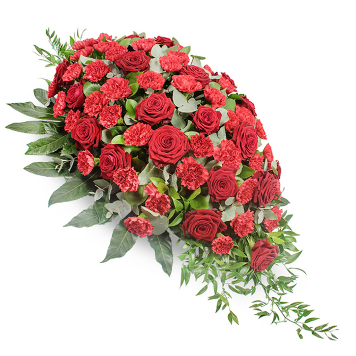 This image shows a large, meticulously arranged red funeral spray displayed on a stark white background, which highlights every contour and colour shift within the flowers. The arrangement is shaped in a classic casket spray style, broad and substantial at the top left, then tapering into a refined point at the lower right, giving it a gentle, cascading motion. At the core sit numerous deep red roses, some fully open, others just beginning to relax their petals. Their velvety texture and rich, wine-red tones create a solemn yet comforting focal point. Nestled between them are clusters of red carnations, slightly brighter in hue, with ruffled, fringed petals that catch the light and add a sense of fullness, much like the textured brick and stonework seen on older Clerkenwell buildings. Surrounding this heart of red blooms is a lavish collar of foliage. Broad, dark green leaves form a steady base, framing the spray and adding depth. Woven delicately throughout are stems of eucalyptus, their rounded, silvery-green leaves softening the intense reds and bringing a slightly smoky, herbal feel to the composition. From the narrowing end of the spray, fine strands of light green trailing foliage extend and curve, enhancing the sense of flow and natural grace. The overall effect is of a thoughtfully designed tribute, the kind of arrangement a Clerkenwell family might select for a funeral at a local church or crematorium, where flowers need to speak gently yet clearly of love, honour, and lasting remembrance.