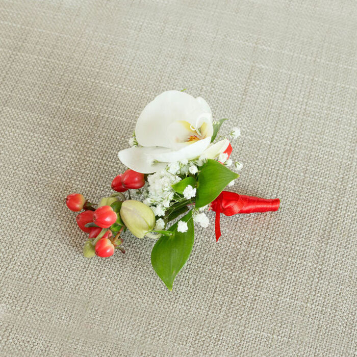 An artfully composed photograph captures a single floral boutonniere placed on a neutral, woven background, the fabric a light beige or oatmeal tone with visible threads that resemble natural linen commonly seen in stylish Clerkenwell studios. In the centre of the arrangement, a pristine white Phalaenopsis orchid unfurls its broad petals, each one smooth, slightly curved, and gently luminous, as though catching a soft, diffused light. At the heart of the orchid, a delicate blend of creamy yellow and soft orange shades gives depth and life to the bloom, offering a subtle hint of warmth in an otherwise cool, elegant palette. Surrounding this focal flower, clusters of tiny white baby's breath blossoms gather like miniature clouds, their fine stems and petite petals adding a feathery, almost ethereal quality that softens the overall look. To one side, a compact cluster of round, glossy red berries introduces a bold accent, their rich, almost cherry-red colour standing out vividly against both the white flowers and the muted background, while nearby, a few paler green berry buds contribute a sense of freshness and growth, reminiscent of seasonal stems sourced from local London markets. Lush green leaves of varying shapes are carefully interspersed among the flowers and berries; some leaves are broader and pointed, others more narrow and elongated, creating an appealing interplay of forms and tones. The stems at the base are tightly wrapped with a vibrant red satin ribbon, its smooth sheen and tidy knot suggesting the skilled hands of an experienced florist. This refined buttonhole feels perfectly suited to weddings in Clerkenwell townhouses, civil ceremonies near Farringdon Station, or winter celebrations across EC1, where a discreet yet polished floral detail can complete a formal ensemble.