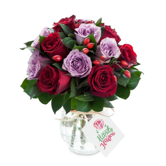 A richly coloured, skillfully composed bouquet centered on an even mix of deep red and soft lavender roses, arranged into a compact, rounded silhouette that reads as both luxurious and approachable. The reds vary from crimson to velvety burgundy; the purples move through lilac and dusty mauve, each bloom fully opened with visible unfurling petals that display their texture and depth. Scattered through the composition are small, bright red-orange rose hips that punctuate the arrangement with sparkles of autumnal colour and a rustic, tactile contrast to the smooth rose petals. Dark green foliage - likely Ruscus or Salal - is woven throughout, its elongated leaves creating pockets of shadow and visual breathing room between the flowers.
The flowers are presented in a bulbous, slightly tapered glass vase with a distinctive mercury or crackled silver finish; its reflective surface softens the light and lends a timeless, antique charm. A simple piece of natural twine is tied around the vase's neck, from which hangs a rectangular white gift tag illustrated with a stylised rose and neat green script, offering a handcrafted, personal touch. Photographed against pristine white, the bouquet's rich tones and the vase's muted gleam are emphasised, suggesting freshness and professional craftsmanship.
Prepared by Clerkenwell Flowers with local sensibility, this arrangement suits romantic gestures, anniversary surprises, or elegant home styling - the kind of bouquet you might pick up after a stroll past Exmouth Market or order for same-day delivery around Clerkenwell and Farringdon when you want something that feels both classic and quietly modern.