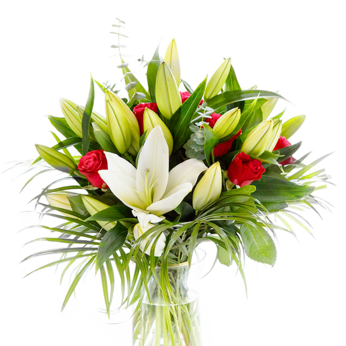 A carefully balanced floral composition centred on a fully open creamy-white oriental lily, its pristine petals curved in a gentle cup to reveal pale yellow stamens and a delicate green pistil. Surrounding this principal bloom are numerous lily buds in shades of lime green and soft yellow, some tightly closed and others beginning to loosen, adding rhythm and anticipation to the arrangement. Scattered artfully between the lilies, rich red rosebuds provide striking punctuation - their velvety, deep-red tones bringing a classic romantic note against the lighter blooms. The greenery is abundant and varied: broad, dark forest-green leaves give structural depth, long, spiky palm-like fronds in a brighter emerald hue cascade outward to introduce movement, and small branches of silvery eucalyptus with rounded leaves soften the palette and add a cool texture. All elements are placed in a clear cylindrical glass vase so that the pale green stems and fresh water are visible, reinforcing the tidy, fresh studio presentation against a bright white background. The image evokes both tactile and olfactory impressions - smooth waxy petals, velvety rose texture, the faint, airy scent of lilies - and reads as an ideal gift for anniversaries, birthdays, or quiet celebrations. As a local Clerkenwell florist I imagine this arrangement delivered to a flat near Farringdon Road or displayed at a tabletop event by St. Clements, crafted with the attention to proportion and freshness customers at Clerkenwell Flowers expect from neighbourhood floristry.