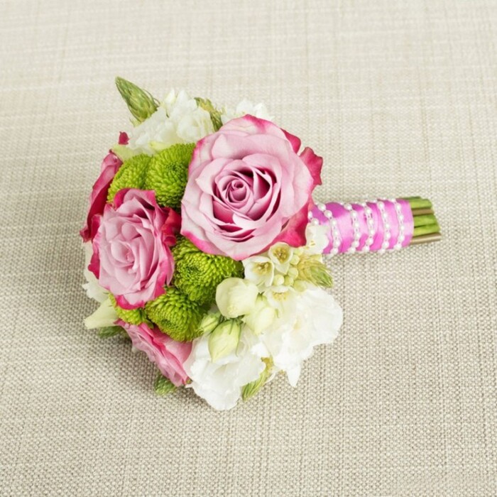 Positioned horizontally on a light tan, subtly woven fabric that resembles fine linen, this compact bouquet looks ready to be lifted and carried down an aisle in one of Clerkenwell's converted warehouse venues. The central focus is a trio of full, open pink roses, each bloom showing off layers of tightly coiled petals that gradually loosen toward the edges. Their colour begins as a soft, warm blush at the heart and deepens into a more vivid, almost magenta tone along the outermost petals, creating an elegant ombré effect that draws the eye. Pressed closely around these roses are rounded clusters of lime-green button chrysanthemums, their many small, neat florets forming firm, cushion-like domes that bring a crisp, contemporary contrast to the rose's romantic curves. Tucked between and around these main blooms are creamy white filler flowers, likely lisianthus or freesia, with multi-layered petals and small buds that add volume and a sense of lightness, like soft clouds floating through the design. Scattered among them are tiny, star-shaped white blossoms with delicate yellow centres, suggesting wax flower or stephanotis, and lending a subtle, intricate charm. The stems at the right-hand side are cut short and tidy, bound together in a tight posy. They are wrapped in a luxurious, bright fuchsia satin ribbon, smooth and slightly reflective, which is then embellished by a slender twist of white faux pearls that crisscross diagonally, giving the handle a jewellery-like finish. Soft, even lighting highlights the colours and textures without casting harsh shadows, allowing the pink, green, and white palette to appear clean and fresh. The entire arrangement feels tailored for weddings, bridesmaids, or engagement celebrations across Clerkenwell, offering a balance of classic romance and modern city style that would photograph beautifully on a table in a local townhouse venue or boutique restaurant.