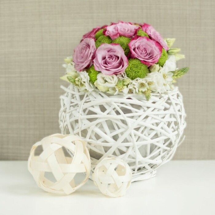 The photograph presents an elegant wedding table centerpiece that feels perfectly at home in one of Clerkenwell's chic venues near Cowcross Street, combining modern design with a soft, romantic heart. Set on a pristine white surface, the focal point is a compact dome of blooms arranged with precise care. The roses, ranging from pale blush to deeper rose-pink and cool lavender, are fully open, their petals layering in spirals that give the bouquet a plush, velvety appearance. Subtle deeper tones at the edges of some petals provide gentle contrast, as though each flower had been brushed with watercolour. Between these roses sit bright green button chrysanthemums, their tight, spherical flower heads adding a bouncy texture and a vivid pop of lime that keeps the arrangement from becoming overly sweet. Tucked at the base and scattered throughout are tiny white filler flowers, each with a star-like shape that softens the transitions between the larger blooms and gives the dome a delicate, almost cloud-like outline. The flowers are cradled in a white, woven vase fashioned from twig-like branches or rattan, formed into a round, openwork globe. Its visible gaps and crisscrossing lines cast slim shadows on the tabletop, enhancing its hand-crafted look. To the lower left of the vase lie two decorative spheres, one medium and one small, made of natural wicker in soft beige tones, repeating the motif of intertwining strands. Behind the arrangement, a gently blurred beige fabric wall suggests linen or burlap, offering a warm, neutral backdrop that lets the pinks, greens, and whites glow. The scene feels serene and welcoming, ideal for a wedding table setting or a stylish celebration in Clerkenwell.