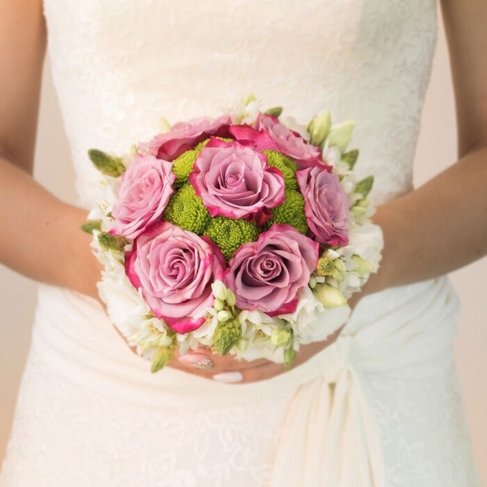 Awash in soft, natural light, a Clerkenwell bride is framed waist-up, her hands gracefully supporting a lush, round bouquet that glows with both colour and promise. The meticulously arranged flowers highlight the expertise of local florists near Exmouth Market. At the centre, full, blooming roses in gentle lavender and antique mauve shift subtly to lively fuchsia at their petal tips, creating a captivating play of colour and texture. Interspersed among the roses and forming a vibrant inner ring, clusters of lime-green button chrysanthemums add a touch of modern freshness, their cheerful hue evoking memories of Clerkenwell's gardens in bloom. The bouquet is bordered by delicate white blossoms-likely modest freesia or petite white roses-interlaced with verdant buds that offer nuanced complexity and depth. The bride's elegantly polished nails, gleaming with pale pink and hints of sparkle, draw attention to her wrists while gently accentuating the arrangement's fragile beauty. Her dress features a breathtaking floral lace overlay along the bodice and sleeves, the intricate patterns echoing the artistry found in the historic architecture of the area. The scene exudes refined bridal charm and a sense of joyful anticipation, illustrating the harmonious blending of colours and textures that make this bouquet not just a focal point of the day, but an enduring emblem of Clerkenwell's vibrant wedding culture.