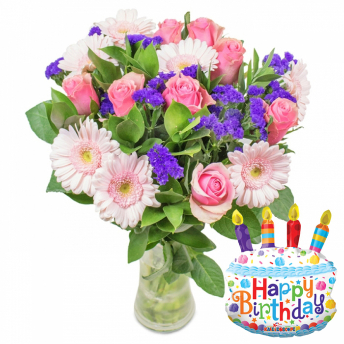 A bright and carefully composed birthday arrangement photographed against a crisp white backdrop, the image pairs a lush glass-vase bouquet with a cheerful metallic cake balloon. On the left, the bouquet is arranged with abundant soft pink roses-petals velvety and fully open-paired with pale pink Gerbera daisies whose layered petals and yellow-green centres lend a classic, sunny presence. Interspersed throughout are clusters of vibrant purple filler flowers, likely statice or limonium, introducing textural contrast and pockets of concentrated colour, while small white spray blossoms add tenderness and fullness. Deep, glossy foliage frames the blooms and supports the rounded silhouette; the stems are visible through clear water in a simple glass vase with a slightly wider base. To the right, a metallic foil balloon shaped like a layered birthday cake stands out: white 'frosting' layers topped with blue piping, a scattering of colourful sprinkles in red, yellow, green and blue, and four multi-coloured striped candles-hints of red, orange, blue and purple-each capped with a bright yellow flame. Across the front the message Happy Birthday is printed in playful letters of orange, green, blue and purple, and a border of stars and confetti decorates the bottom edge; a small 'KALEIDOSCOPE' logo is visible along the lower rim of the balloon. The ensemble conveys a joyful celebration, the precise handiwork of a local florist who might be supplying birthday gifts for colleagues near Charterhouse Square or visiting customers from Smithfield Market.