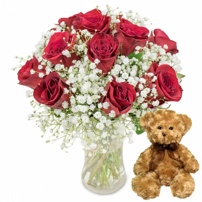 This carefully lit studio photograph showcases a classic romantic gift set that could easily be imagined en route to a Clerkenwell loft close to St John's Square. On the left, a luxurious bouquet of roughly twelve deep red roses fills a clear glass vase, the blooms arranged to create a full, rounded silhouette. Each rose is a study in velvety texture and depth of colour, ranging from rich, wine-red tones at the outer petals to darker, shadowed crimsons at the heart of each flower. The petals are densely layered, some slightly cupped, others fully open, suggesting freshness and meticulous handling. Interlaced among the roses is an abundance of white baby's breath, its countless petite blossoms forming an airy lace that brightens the composition and adds volume without overwhelming the roses. Through the transparent sides of the vase, straight green stems stand in clear water, angled with the quiet precision of a professional florist from Clerkenwell Flowers, who understands balance and proportion. To the right of the vase, a medium-sized teddy bear sits comfortably upright. Its light brown plush fur has a soft, almost downy texture, subtly highlighted by the even white lighting. Rounded paws rest naturally on the surface, while small black button eyes gaze forward with a gentle, friendly expression. A slim dark brown satin ribbon, tied neatly at the neck, provides a simple yet refined detail. The white background, free of distraction, enhances the sense that this is a thoughtfully curated gift, ideal for a surprise anniversary delivery to an office near Farringdon Station, a Valentine's Day treat for a partner living off Clerkenwell Road, or a tender birthday gesture filled with warmth and affection.