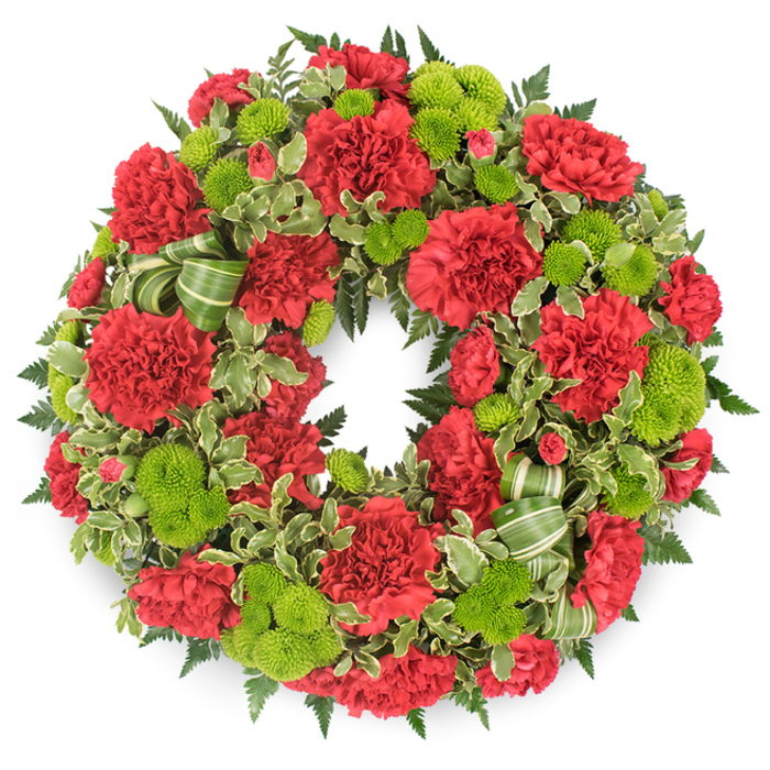 Against a stark white background, a meticulously crafted circular funeral wreath radiates with the rich contrast of dark red and bright lime green, each flower placed with the precision and care you might expect from an experienced Clerkenwell florist. The ring is packed tightly with deep red carnations whose multi-layered, frilled petals form soft, undulating surfaces that catch the light and give the wreath a plush volume. These crimson blooms are thoughtfully spaced with clusters of lime green button chrysanthemums, their perfectly spherical heads composed of countless tiny florets that create a smooth, almost velvety texture. Together, the two flower types form a rhythmic pattern of colour around the circle, alternating between depth and brightness. Surrounding and supporting the flowers is an abundant mixture of foliage: dark green fern fronds fan gently outward, adding a feathery edge, while variegated pittosporum brings a lighter note with leaves that show creamy white margins around deep green centres. Nestled discreetly among the greenery is a decorative ribbon in soft and darker green stripes, looped and tucked so that it mirrors the round form without distracting from the blooms. The lighting is even and crisp, allowing every shade of red and green to stand out, while a faint shadow beneath the wreath gives it a subtle three-dimensional presence. This tribute feels composed for a thoughtful farewell in the Clerkenwell area, suitable for services near Farringdon, quiet chapels tucked off Rosebery Avenue, or memorials held in local community spaces where families gather to honour a loved one with dignity and warmth.