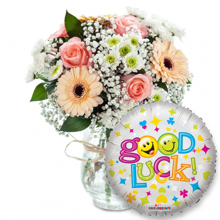 Lovingly composed and captured on a white field, this encouraging 'Good Luck!' gift pairs fresh-cut florals with a playful foil balloon, the sort of arrangement Clerkenwell Flowers might prepare for a neighbour on the eve of an important day. The bouquet, set left of centre in a clear glass vase tied with rustic twine, blends warm peach Gerbera daisies with their deep brown centres, delicate light-pink roses whose petals fold in soft spirals, clouds of white baby's breath that soften the silhouette, and clusters of small white and lime-green chrysanthemum florets for a crisp accent. Rich green foliage slips between stems, creating pockets of shadow and structure; the tactile contrast is clear - silky rose petals, slightly textured Gerbera heads, and the papery snap of chrysanthemum florets - while the scent remains gentle and uplifting. In the foreground, slightly overlapping the base of the vase to the right, a round metallic balloon glows with a silver-white sheen and is scattered with colourful hearts, stars and confetti shapes. Bold, bubbly letters spell out Good Luck! across its face in multiple bright hues; the two 'o's in Good are replaced by cheerful yellow smiley faces, one winking, the other sticking its tongue out, lending a warm, mischievous charm. A small Kaleidoscope label is visible at the balloon's rim. The overall impression is intimate and optimistic - a feel-good ensemble perfect for exam send-offs, interview support or a celebratory new chapter - available for local delivery across Clerkenwell, Exmouth Market and nearby Hatton Garden.