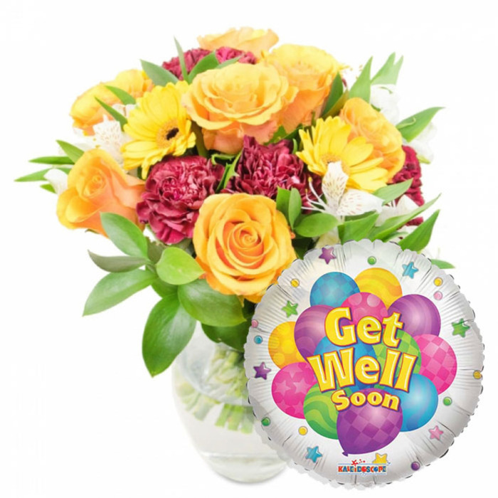 A cheerful, carefully curated Get Well Soon gift ensemble that feels like something we might prepare at Clerkenwell Flowers for a patient at a nearby ward or a friend resting near Leather Lane. The main element is a full, lush bouquet arranged in a clear glass vase; the stems plunge into visible water, and the vase's transparency gives an honest, unfussy look. Orange and warm yellow roses open at their centers, petals layered like soft, sunlit spirals and emitting a mild citrus-honey scent that soothes rather than overwhelms. Bright yellow gerbera daisies lift the arrangement with wide, cheerful faces, while deep maroon or burgundy carnations add a rich, textured counterpoint; their ruffled edges create visual depth and a comforting density. Sprays of small white alstroemeria peek between the larger blooms, lending delicate contrast and tiny flecks of pattern. Varied green foliage-glossy leaves and slender, airy fronds-frames the bouquet, giving it fullness and a freshly-gathered-from-the-market feel. Positioned to the lower right and overlapping the bouquet slightly, a round metallic silver Mylar balloon adds a celebratory accent: its reflective surface holds a whimsical image of multicolored cartoon balloons in blue, purple, pink, yellow and green, some decorated with stripes, polka dots or checkerboard patterns. Across the center a friendly message in yellow-and-blue lettering with a soft shadow reads Get Well Soon, while small starbursts around the white border increase the festive tone; a small KALEIDOSCOPE logo is visible at the balloon's bottom edge. This pairing of bright blooms and a playful foil balloon communicates care and hopeful encouragement-an ideal way to send comfort locally within Clerkenwell, St John's Square and the surrounding streets.