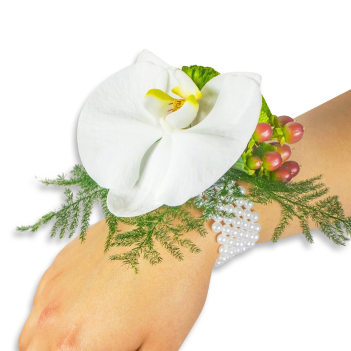 Displayed on a light-skinned wrist against a spotless white backdrop, this elegant wrist corsage combines classic floristry with a contemporary eye for texture and colour. Its centerpiece is a solitary white Phalaenopsis orchid, broad and smooth as porcelain, the petals arranged in a sculpted, open form that feels poised and refined. At the heart of the orchid, the labellum presents a concentrated yellow with warmer orange hints, a small but decisive note of colour that anchors the composition. A generous bed of asparagus fern beneath the bloom provides a lush, feathery base in bright green, the fine foliage creating depth and a whispery contrast to the orchid's solidity. To the upper right, a compact cluster of Hypericum berries contributes tactile contrast and seasonal warmth, their hues graduating from pale green through rosy tones to a deep red-crimson that reads almost like tiny garnets. A ruffled lime-green accent leaf nearby adds another layer of texture and a fresh highlight. The arrangement is secured on a refined multi-strand pearl bracelet - numerous small, lustrous white beads that circle the wrist comfortably and lend a timeless, almost heirloom quality. Photographed in soft, even light, the corsage reveals its textures clearly: the orchid's satiny surface, the fern's filigree, the berries' sheen, and the pearls' soft glow. Crafted with steady hands and an eye for balanced scale, this corsage-prepared by local florists familiar with Clerkenwell events-is ideal for weddings, proms, or any formal occasion where understated elegance is desired.