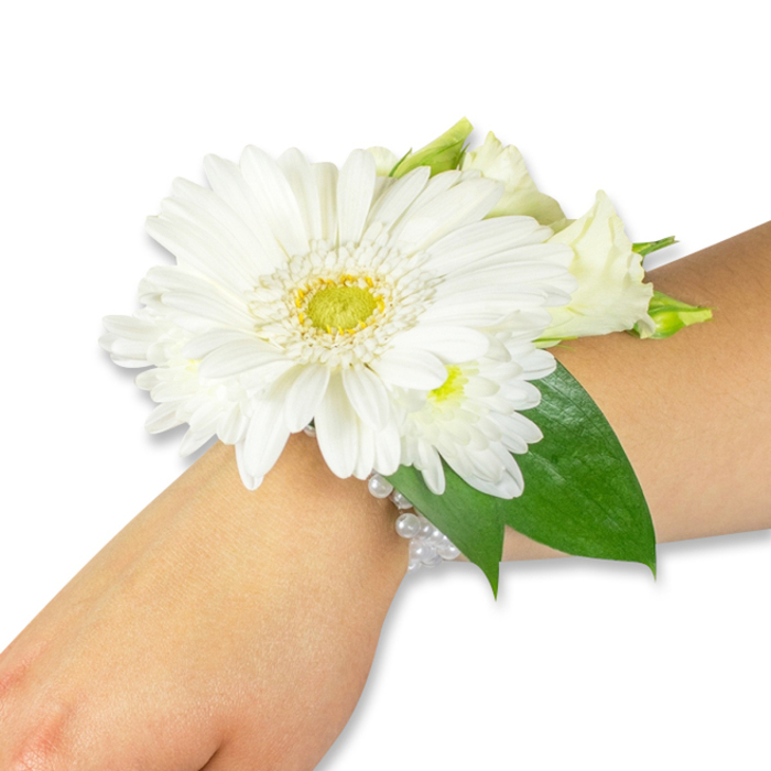 An elegant white floral wrist corsage is artfully arranged along a fair-toned forearm, photographed against a pure white background to showcase every curve and surface. The centerpiece is a pristine white Gerbera, its broad, velvety petals radiating outward from a textured yellow-green disc that adds a focused point of brightness. Surrounding that prominent bloom are clusters of neat, white spray chrysanthemums and creamy lisianthus buds and open petals, layered to create soft volume and a nuanced palette of whites. Behind the flowers, deep green salal or ruscus leaves provide a structured, verdant backdrop; their glossy surfaces and firm veins anchor the composition and give the arrangement a pleasing depth. The entire floral cluster is secured to the wrist by a refined multi-strand bracelet of tiny white pearls, each bead reflecting light with a mother-of-pearl glow and lending a touch of vintage romance to the contemporary simplicity of the blooms. The imagery evokes the gentle scent of fresh-cut flowers-green, cool, and slightly herbaceous-and the tactile contrast between silky petals and smooth beads. Designed for formal occasions such as bridal parties, proms or graduation ceremonies, this corsage is the sort of piece a local Clerkenwell florist would prepare with care for a walk down St John Street or a photograph outside Farringdon Station. Crafted with balance and restraint, this wearable bouquet reads as an emblem of purity and celebration while remaining comfortably rooted in the local streets and quiet moments of the neighbourhood. Clerkenwell Flowers prepares similar designs for same-day delivery across the area when freshness matters most.