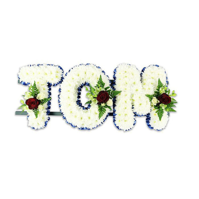 A carefully made memorial piece spells out the name TOM in striking three-dimensional letters, each face covered with an even, dense layering of white chrysanthemum blooms that create a plush, tactile surface. Each letter's perimeter is neatly bordered with a royal blue ribbon that gives reassuring structure and a sharp, elegant contrast to the snow-white flowers. Centered within the lower portion of each letter sits a composed spray: a single, deep red rose anchors the cluster, its velvety petals symbolising enduring love and respect. Surrounding the red focal are smaller cream-colored spray roses and tiny white buds, while slender fern fronds and other lush greens weave through the composition to supply texture, shadow and graceful movement. The letters are supported by a narrow dark metal bar running subtly across the rear base, indicating the arrangement is freestanding for display in a church, funeral home or a graveside service. Photographed against a pure white background, the design's colours - stark white, regal blue and somber red - are allowed to read clearly and solemnly; delicate shadows enhance the sense of volume and craftsmanship. This tribute feels composed and intimate, something a Clerkenwell florist might prepare for a wake near St John's Street or for delivery to a memorial near Exmouth Market, created to convey sympathy and dignified remembrance through considered floral artistry.