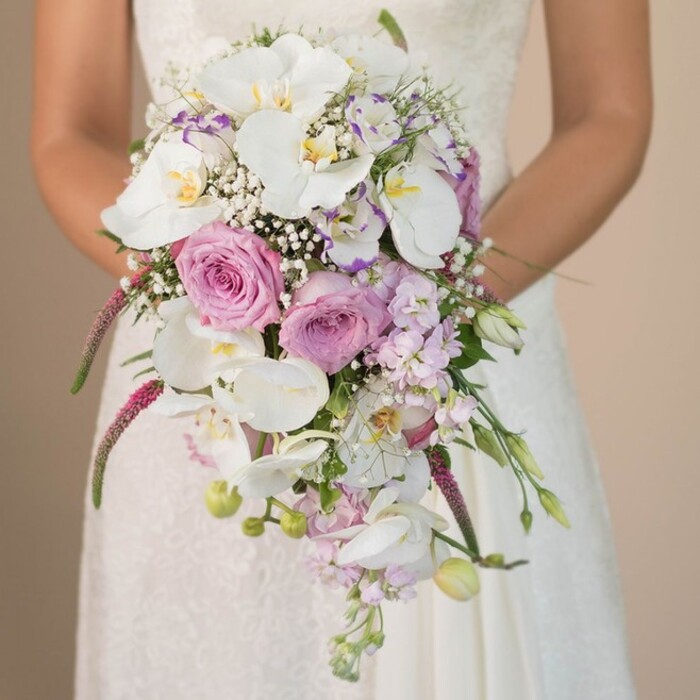 Captured in an elegant moment against a serene, neutral background, a Clerkenwell bride holds a lavish cascading bouquet, its artistry rivaling the community's renowned creative ethos. The arrangement unfurls from her hands in a gentle waterfall, with layers of white phalaenopsis orchids at the center-each broad petal reflecting daylight with a subtle pearlescence and highlighted by soft yellow throats. Interwoven are clusters of blush and lavender roses, their petals tightly swirled and rich with natural scent, echoing the local rose blooms of Spa Fields in early summer. Infusions of gypsophila (baby's breath) interlace the bouquet, creating an ethereal, cloudlike texture that complements soft lavender and lilac blossoms, perhaps stock or sweet pea, nodding through the bouquet with delicate whimsy. Cascading magenta amaranthus threads downwards, their plush, trailing tendrils adding textural interest and movement-reminiscent of Clerkenwell's artistic vibrancy. Polished green leaves and a scattering of closed buds complete the arrangement, suggesting ongoing growth and renewal, important themes for any wedding day. The delicate, understated lace along the bride's bodice peeks from beneath the florals, harmonizing the softness of her attire with the bouquet's romantic hues. Designed for celebrations in Clerkenwell's unique venues, this bouquet exudes both sophistication and freshness-a timeless choice for couples seeking floral elegance rooted in local tradition and flair.
