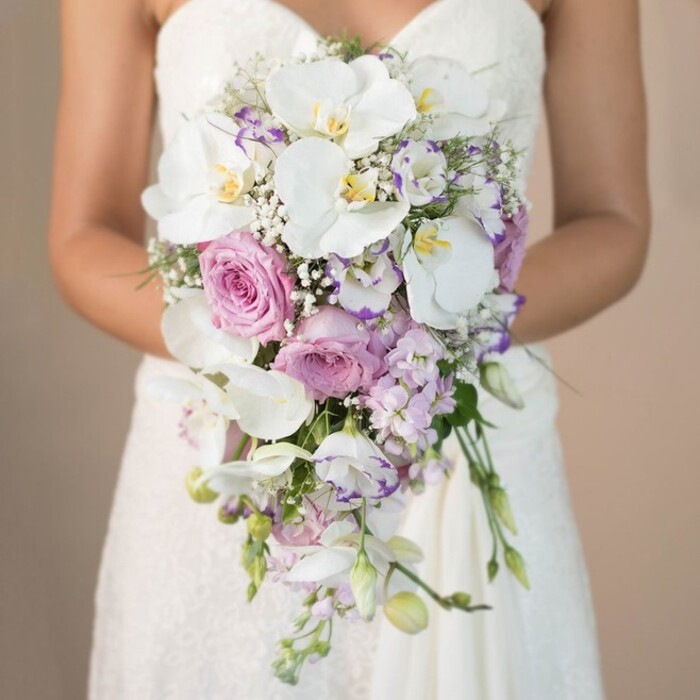 A stunning close-up unfolds in the glowing heart of Clerkenwell: a bride, visible from the chest down, cradles a lavish, cascading bouquet-a vivid embodiment of local wedding elegance. In this sophisticated arrangement, glossy white phalaenopsis orchids sweep gracefully downward, their broad petals and subtle yellow throats reflecting the chic style synonymous with Clerkenwell designers. Nestled among them, sumptuous soft pink garden roses unveil their tightly layered petals, while smaller sprigs of lavender and pale purple stock flowers ripple with gentle movement and texture. White lisianthus, the ruffled edges sometimes blushed deep violet, mingle among the blooms to introduce yet another note of luxury. The artistry is further heightened by dainty clusters of baby's breath and draping green leaves, all woven to maintain a light, voluminous silhouette. The bride's strapless lace gown, embroidered with floral patterns, creates a visual link between fabric and flowers, both resonating under the softly diffused light common to Clerkenwell studios. Her arms encircle the bouquet with a quiet sense of anticipation, directing every attention to this masterpiece. This image is an invitation-to imagine the aroma of fresh petals drifting toward Farringdon Road, to feel the care of Florists in Clerkenwell, and to appreciate a bouquet perfect for contemporary or classic wedding celebrations across EC1.
