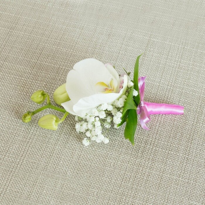 A single, carefully composed orchid boutonniere is laid out horizontally across a softly woven, light beige linen surface, the texture of the fabric subtly visible beneath the stems and petals, like the gentle weave of a handmade table runner in a Clerkenwell apartment. The main bloom is a pristine white Phalaenopsis orchid, its wide, slightly arching petals smooth and unblemished, creating an almost sculptural form. At the centre of the orchid, the throat shows delicate detailing in soft yellow and the faintest veil of pale pink, adding warmth and depth without disturbing the flower's overall purity. Encircling the base of the orchid, small clusters of snow-white baby's breath form a cushion of airy blossoms; their numerous, bead-like flowers sit on fine, branching stems, giving an impression of lightness and romance. Just behind and among the blooms, a few slim, vibrant green leaves emerge, their fresh colour hinting at the living plant from which this careful cutting was taken. To one side of the arrangement, a short offshoot bears two lime-green orchid buds, smooth, closed, and gently tapered, suggesting petals still tucked safely within, poised to open after the festivities. The stems of the orchid, gypsophila, and foliage are gathered neatly and wrapped with a bright fuchsia pink satin ribbon, tied into a small, precise bow with short tails that rest along the fabric. The ribbon's glossy surface catches the soft lighting, introducing a joyful, contemporary splash of colour that contrasts beautifully with the neutral background and white petals. This boutonniere feels ideal for a groom's jacket or a sophisticated corsage at a wedding reception held in one of Clerkenwell's converted warehouse venues or near Spa Fields, embodying a balance of modern style and timeless elegance suitable for proms, civil ceremonies, and formal city celebrations.
