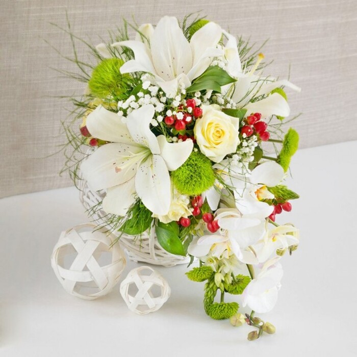A sophisticated white and green floral centerpiece is showcased on a pristine white surface, set against a subtly textured, warm beige background that feels calm and understated, like the interior of a Clerkenwell design studio. The flowers are arranged in a low, round basket crafted from white woven wicker, its interlaced strands forming a gentle, organic base for the display. Rising from this base, several large Oriental lilies in pure white dominate the upper portion of the design, their open trumpet-shaped blooms revealing delicate spots and prominent stamens, giving a sense of fragrance and luxury. Slightly off-centre at the top, a single ivory-cream rose unfolds in layered petals, creating a romantic focal point that draws the gaze inward. Around the central flowers, a lush tapestry of greenery includes feathery fronds, slim blades of ornamental grass, and broader, darker leaves, all contributing movement and depth. Rounded green pom-pom blooms, likely dianthus or Santini chrysanthemums, punctuate the foliage, their soft, moss-like texture adding interest and volume. Threaded delicately between these elements, tiny white baby's breath blossoms form hazy clusters, while glossy red hypericum berries provide joyful bursts of colour, reminiscent of seasonal celebrations around nearby Farringdon and Smithfield. Flowing from the right-hand side, a graceful curtain of white phalaenopsis orchids tumbles down in a cascade, their pristine petals and slender stems giving the design a bridal, waterfall quality that would suit a wedding breakfast near Clerkenwell Green. To the left, two decorative white lattice spheres sit on the table, one slightly larger than the other, echoing the woven basket and completing the composition. The arrangement radiates understated opulence and timeless elegance, ideal for wedding tables, upscale events, or refined home décor in central London.