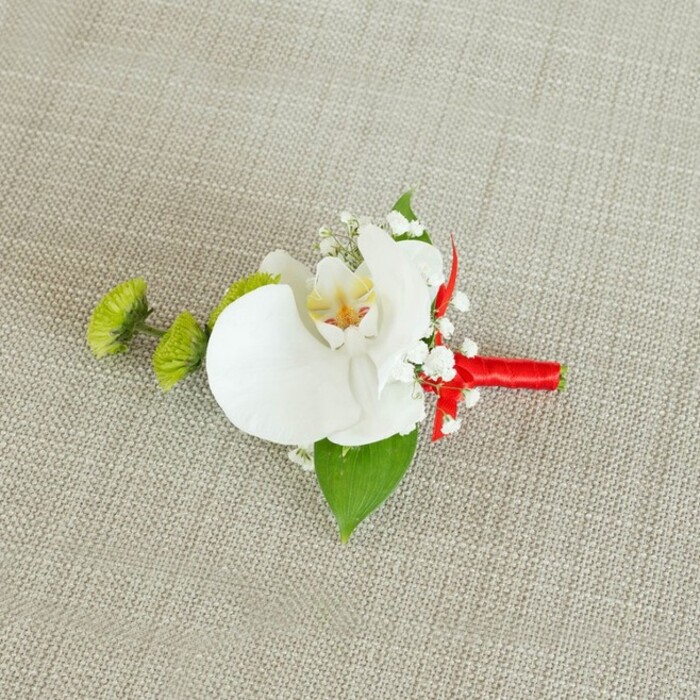 A refined orchid boutonniere is displayed flat against a light beige, linen-textured backdrop, its soft, natural weave suggesting the relaxed elegance of a Clerkenwell loft or intimate town hall ceremony. At the centre of this floral piece is a large white Phalaenopsis orchid bloom, its petals broad, smooth, and gently undulating, forming a graceful, butterfly-like shape. The orchid's throat is delicately accented with a warm wash of pale yellow that transitions into faint reddish-orange or light brown markings, offering a subtle depth of colour that enriches the otherwise pure white flower. Encircling this central blossom, clusters of airy white baby's breath cluster like fine lacework, each tiny bloom adding a sense of lightness and romance, as if a soft mist had settled around the orchid. To the left side, two vibrant lime-green, pom-pom-shaped chrysanthemum buds-or similarly textured blooms-provide a striking contrast, their compact, spiky petals introducing playful structure and a contemporary twist that feels at home in the creative streets off Farringdon Road. Beneath the flowers, a broad, deep green leaf extends as a natural base, while a smaller, darker leaf peeks out closer to the stems, ensuring the design feels anchored and balanced. The stems are tightly bound with a rich crimson satin ribbon, wrapped in smooth, precise coils and finished with a neat, decorative knot that contributes a bold splash of celebratory red. The consistent, slightly coarse linen background allows every petal, bead of colour, and strand of baby's breath to stand out clearly. This carefully composed boutonniere evokes the anticipation of a wedding day in Clerkenwell, ideal for a groom, best man, or guest who appreciates understated luxury and modern floral design.