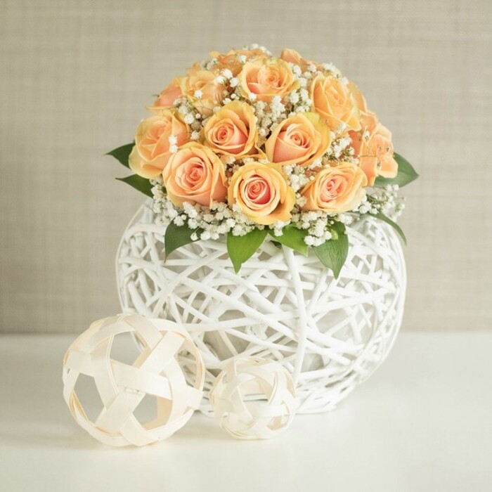 The image presents a refined floral centerpiece, where approximately two dozen peach and apricot roses are arranged in a perfectly domed bouquet. Each rose is at a slightly different stage of opening, some still cupped with tightly furled hearts, others more generous and layered, revealing intricate spirals of petals. The colours blend from deeper, almost honeyed orange at the centre of each bloom to softer, creamy peach at the outer edges, creating a gentle, luminous effect that feels both romantic and contemporary. Interwoven among the roses are clusters of delicate white gypsophila, or baby's breath, their tiny star-shaped blossoms forming airy clouds that break up the solid mass of colour and introduce a sense of breath and space. Hints of rich green foliage peek from beneath the flowers, suggesting a hand-crafted arrangement made with fresh stems, like one prepared early in the morning for a Clerkenwell apartment overlooking St John Street. The bouquet is nestled into a striking, spherical vase formed from thin white strands woven into a complex lattice, the open structure allowing you to glimpse the interior while offering a sculptural, almost architectural presence. This vase sits on a smooth, bright white surface, likely a tabletop or sideboard, where two additional decorative orbs are placed in front. These smaller spheres, crafted from light, natural-toned material, echo the same woven, geometric texture at different scales, adding visual rhythm and a touch of playful design. Behind it all, a soft beige or off-white wall with fine vertical texture serves as a clean, unobtrusive backdrop. Gentle, even lighting from the side highlights the velvety petals, the intricacy of the weaving, and the subtle shadows that bring depth, making the scene ideal for illustrating sophisticated wedding décor, home staging in central London, or a carefully styled arrangement from a trusted local florist.