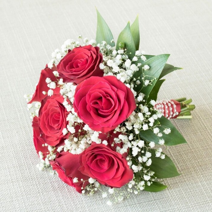 This exquisitely styled bouquet, arranged by Clerkenwell Flowers, captures the essence of sophisticated romance found along the storied streets of Clerkenwell. A cluster of five or six plush, fully opened red roses sit at the heart of the design, their velvety petals radiating a rich, deep red that conjures feelings of passion and enduring love-ideal for a local wedding at one of the area's historic venues. Intertwined with the roses are generous sprays of tiny white baby's breath (Gypsophila), whose airy, cloud-like presence offers a gentle, softening contrast against the bouquet's intense central blooms. Fresh, emerald-green foliage, possibly ruscus or salal, adds both depth and dimension, evoking the serene greenery of quiet Clerkenwell gardens tucked away behind Georgian terraced houses. The finishing detail, a length of vibrant red and white striped ribbon coiled precisely around the stems with a strand of petite pearls, evokes timeless bridal elegance while adding subtle texture and sparkle. The entire bouquet is gently placed on a pale, textured backdrop-linen or hessian-allowing its vivid hues and careful craftsmanship to stand out. The composition is bathed in soft, natural light, celebrating each floral nuance and intricate detail. This bouquet invites the senses to imagine the subtle perfume of fresh blooms mingling with the anticipation of a joyful celebration, crafted with care for anniversaries, romantic gifts, or heartfelt moments in EC1.