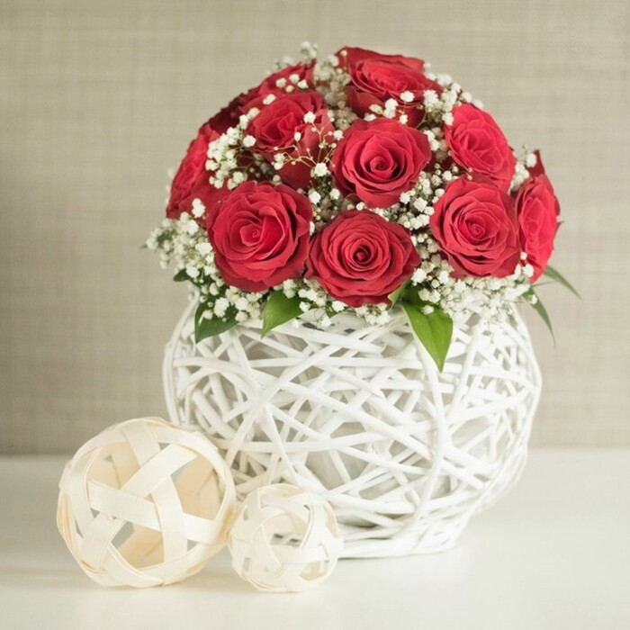 This image presents an elegant floral display that would feel at home on a wedding table in one of Clerkenwell's intimate restaurants or historic venues. At its centre is a generous, spherical bouquet of deep red roses, their petals velvety and full, spiralling tightly to create a sense of depth and luxury. The blooms sit closely shoulder to shoulder, forming a rounded dome that speaks of romance and devotion, like a classic love story rendered in petals. Interwoven between the roses are airy stems of white baby's breath, each tiny flower a delicate dot of brightness that softens the intensity of the red and gives the arrangement an almost cloud-like aura. Small glimpses of fresh green leaves peek out, adding a natural rhythm and helping the bouquet feel freshly gathered. The flowers are presented in a distinctive round white vase with an open-lattice structure, resembling white wicker or rattan strands woven into a sculptural nest. The spaces between the strands allow light to pass through, creating interesting shadows on the surface below and reinforcing the modern, handcrafted character one might associate with Clerkenwell Flowers. To the left of the vase, two decorative spheres crafted from pale, flat woven strips sit side by side, the larger sphere nearer the viewer and a smaller one just behind. Their off-white and cream tones harmonise with the vase while adding a playful variation in size. All of this rests on a smooth, clean white tabletop, set against a softly textured neutral wall that fades gently into the background. The scene feels calm, curated, and romantic-a perfect visual for showcasing wedding reception flowers, anniversary centrepieces, or refined home décor pieces available for local delivery around Clerkenwell.