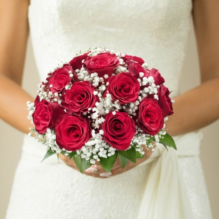 Captured against the understated elegance of a Clerkenwell bridal suite, this evocative image centres on a close-up of a bride holding a classically styled round bouquet. Vibrant, full-petaled red roses form the heart of the arrangement-some budding tightly, others lush and open-offering a textural journey across each velvety bloom. Interspersed with artistic care are abundant tufts of snowy white baby's breath, whose dainty texture lifts the deep reds into a light, cloud-like symphony. A scatter of glossy green leaves tucks beneath the florals, adding a fresh accent reminiscent of the leafy avenues winding through Clerkenwell's Exmouth Market. The bride's subtly manicured hands anchor the bouquet, while the intricately patterned wedding gown-suggesting lace or delicate embroidery-layers further refinement onto the scene. A flowing white sash cascades down the right, hinting at movement and a gentle breeze, perhaps like those that drift through Farringdon on a wedding morning. Bathed in soft, natural light, the imagery blurs gently into a creamy background, isolating the bouquet and bride as the pure focus of the celebration. This composition, designed by Clerkenwell Flowers, captures not just expert floristry but the serene joy and sophistication that local couples seek for their special day in the heart of London.