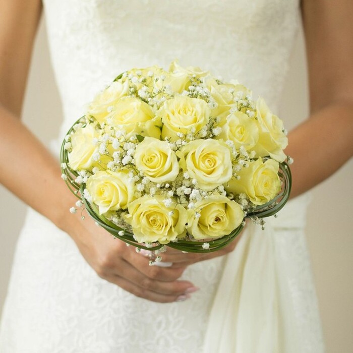 A beautifully composed photograph draws the eye to the hands of a bride-seen from the chest down, the creamy white of her wedding gown subtly reflecting the morning light just off Farringdon Road. She cradles a truly exquisite bouquet: dozens of pale yellow roses, each at the height of bloom, create a lush mound of soft petals, their golden tones glowing against the dress's delicate texture. Intertwined among the roses are sprays of ethereal white baby's breath, bringing a sense of airiness and classic elegance that harks back to Clerkenwell's Victorian garden courtyards. Framing the base, slim bands of dark green foliage or intricately twisted wire offer both support and a stylish contrast, reminiscent of local artisanal touches. The bride's hands, perfectly manicured in soft nude lacquer, emphasize tenderness and anticipation as they hold the bouquet aloft. The gentle blur of the background ensures that both bouquet and wedding attire stand as the unmistakable centrepiece-icons of celebration, artistry, and hope. Every detail, from the fresh-cut roses to the careful placement of each baby's breath stem, tells a story rooted in Clerkenwell's renowned floral tradition, making this arrangement a perfect tribute to love and refined taste.