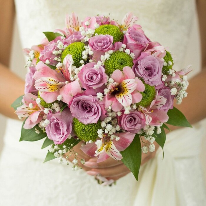 A radiant, spherical bridal bouquet glows in a bride's hands, poised against the delicate lace of a wedding gown-capturing a moment that feels quintessentially Clerkenwell, perhaps just steps from the creative energy of Exmouth Market. The composition is a masterful blend: central to the display are roses in soft pastel pinks and muted lavender, their petals layered in gentle openness, some glistening faintly as though touched by a morning mist from nearby Spa Fields. Peppered throughout are bold, lime green button chrysanthemums, the roundness and zest of their form offering a vibrant, modern counterpoint to the traditional roses. Ribbons of pink alstroemeria, with intricate markings of maroon and yellow, add a sophisticated air and hint at the international flair that defines Clerkenwell life. Fine clouds of white gypsophila fill the bouquet, their airy texture amplifying the sense of lightness and romance. Deep green rose leaves line the base, framing the array with organic elegance. The bride's hands-one adorned with a simple wedding band-grasp the carefully wrapped stems, the bouquet's palette echoing the fresh, artistic vibe found in local galleries. This floral creation feels elegantly curated for couples seeking a blend of tradition and trend, perfect for a spring or summer wedding reception at the heart of Clerkenwell's vibrant community.
