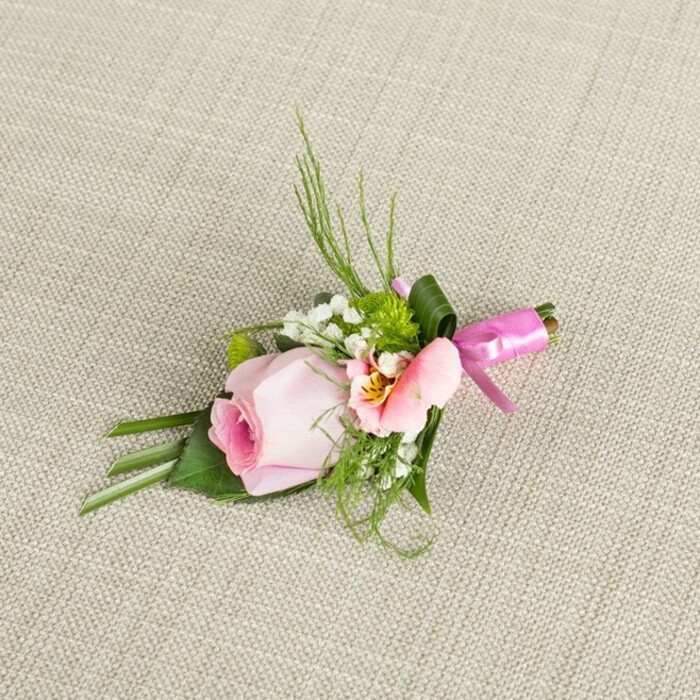 Positioned diagonally on a softly textured, oat-toned fabric suggestive of the high-quality linens found in Clerkenwell's boutique studios, this elegant boutonniere weaves together the area's love of artistry and tradition. A central soft pink rose, its petals tightly coiled, radiates freshness and hope, complemented by a lighter peach-pink alstroemeria whose open petals hint at warm yellow near the centre and subtle flecks of darker pigment. Sprigs of cloud-like baby's breath lend airiness, while bright green mini mums add a striking touch of texture reminiscent of the foliage in nearby Percy Circus. At the base, an array of foliage-from slender, upright blades to broad, folded leaves-creates depth and structure, grounding the arrangement much like the sturdy facades of Clerkenwell's historic lanes. A wide, deep green leaf is methodically wrapped around the bound stems, adding polish, while the base is finished with a vibrant, fuchsia satin ribbon tied into an elegant bow. This handcrafted accent, glowing softly in even daylight, is ideal for a wedding, prom, or distinguished gathering at local venues, the perfect touch for a lapel or wrist. It embodies the intersection of timeless design and Clerkenwell's modern vibrancy, capturing a sense of sophistication and celebration in every petal and ribbon gleam.