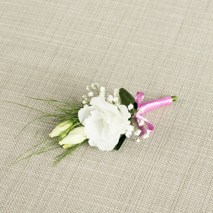 An elegant boutonnière is displayed on a diagonally aligned swathe of light beige, textured fabric, the weave resembling the kind of natural linen often used in stylish Clerkenwell interiors. The arrangement is centred around a single, fully open white lisianthus flower, its numerous ruffled petals curling gently outwards in layered, creamy folds that create a soft, rounded form. Each petal edge is subtly defined under the soft, even lighting, giving the bloom a velvety, almost luminous appearance. Nestled slightly below and to the left of this main flower are two slender lisianthus buds, their elongated shapes tinted with pale green and cream, capturing that quiet, in-between moment before they unfurl. Fine stems of white gypsophila weave through the design, their tiny blossoms clustered like miniature stars, adding a delicate, cloud-like fullness around the focal flower. From behind the central bloom, narrow, grass-like blades of dark green foliage sweep out towards the lower left corner, introducing gentle movement and a sense of natural flow, while one broader, rich green leaf is tucked at the back, providing structure and a deeper contrast. The stems converge neatly at the lower right, where they are wrapped in a band of vivid fuchsia satin ribbon, tied into a small, precise bow. The ribbon's bright pink hue and soft sheen stand out against the calm background and the fresh white and green tones, offering a modern, playful twist on a classic wedding accessory. This detailed close-up could easily be a groom's buttonhole ready for a ceremony in a Clerkenwell gallery or intimate registry office, its careful craftsmanship and fresh textures capturing the romance and anticipation of the day.