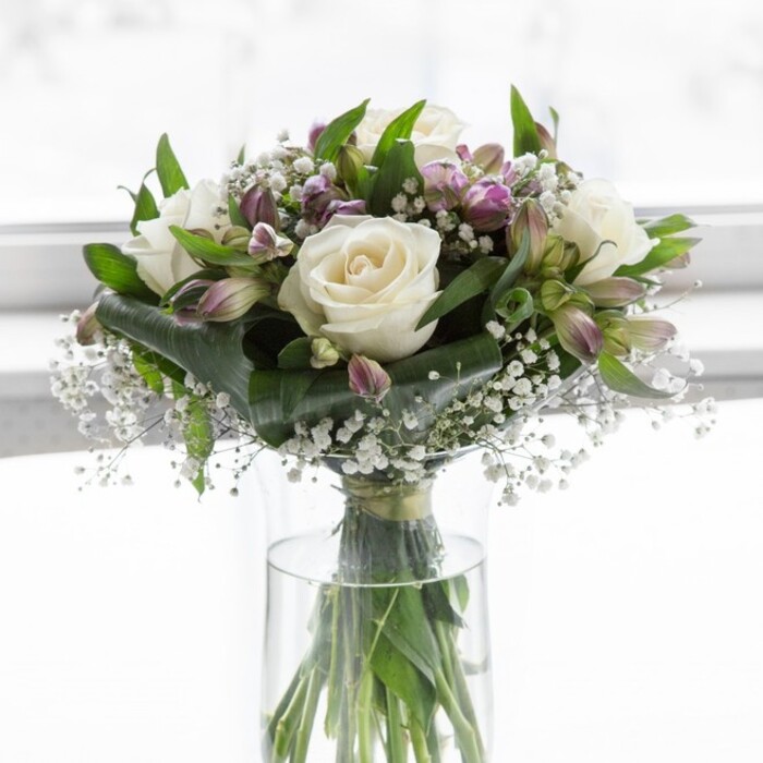 In this image, an elegantly composed bouquet of white and lavender blooms rests in a simple, clear glass cylinder vase, positioned against a luminously soft background that suggests daylight streaming through a Clerkenwell studio or apartment window. The focal flowers are creamy white roses, fully opened with generous, spiralling petals that create a plush, romantic centre. They are interwoven with numerous stems of lavender and muted purple alstroemeria, some still enclosed in their protective green sepals and others just beginning to open, revealing slender petals with fine, darker veining that adds depth and character. Tiny white blossoms of baby's breath scatter delicately among the roses and alstroemeria, their airy clusters giving the impression of a light mist drifting through the arrangement. Rich green foliage in multiple shapes-broad, dark leaves and more slender, elongated greenery-frames the flowers and gently rounds the bouquet, providing both contrast and structure. Through the transparent vase, the cleanly cut stems are visible, fanning out in clear water, a quiet nod to the technical care and conditioning behind the design. The lighting is natural and even, giving the whites a creamy warmth and the purples a gentle, almost twilight softness, reminiscent of a calm evening walk past the historic streets and brick facades around Clerkenwell Green. The overall mood is one of understated elegance and peace, suggesting a bouquet that might be delivered locally to mark a milestone birthday, to offer sympathy without heaviness, or to bring a sense of calm beauty to a home office overlooking Hatton Garden or Farringdon. It feels like a considered piece of floral artistry, composed with patience and a light touch.