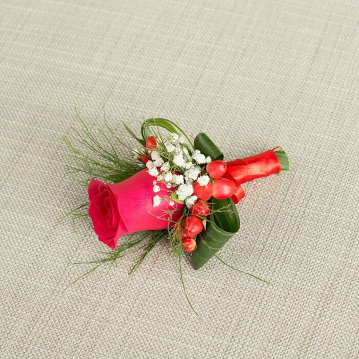 Set against a softly woven, light beige fabric that recalls the understated interiors of Clerkenwell's converted warehouses, this boutonniere is photographed from a slightly elevated angle, letting every flourish of floral craftsmanship be clearly seen. The central flower is a bold fuchsia pink rose, its velvety petals layered closely together, spiralling outward in a saturated whirl of colour that suggests passion and joyful anticipation. The rose is in full, fresh bloom-open enough to show depth and structure, yet firm and unblemished, with just the hint of a sweet fragrance you can almost imagine. Encircling its base are fine, cloud-like clusters of white baby's breath, the tiny blossoms gathered in sprays that soften the composition and catch the light like little drops of cream against the bright pink. Interspersed among these delicate whites sit glossy red hypericum berries, their round forms adding a playful, rhythmic pattern and a deeper note of red that bridges the gap between the rose and the ribbon binding the stems. Around and beneath the flowers, a combination of foliage brings structure and movement: some leaves are broad and dark, expertly curled to cradle the rose, while slender, feathery greenery stretches outward in wisps, echoing the natural looseness of garden growth even in this compact design. The stems are bound tightly together with a luxurious red satin ribbon, wrapped smoothly and finished in a small, precise knot or bow, giving the piece a polished, ceremonial feel. Soft, even lighting falls over the scene, highlighting the textures of the fabric background, the gleam of the berries, and the subtle sheen of the ribbon. This carefully composed floral accent could easily be pinned to a lapel for a wedding at a modern venue off Farringdon Road or worn at a civil ceremony near Clerkenwell Green, its colours and textures marking the wearer as part of a cherished, shared celebration.