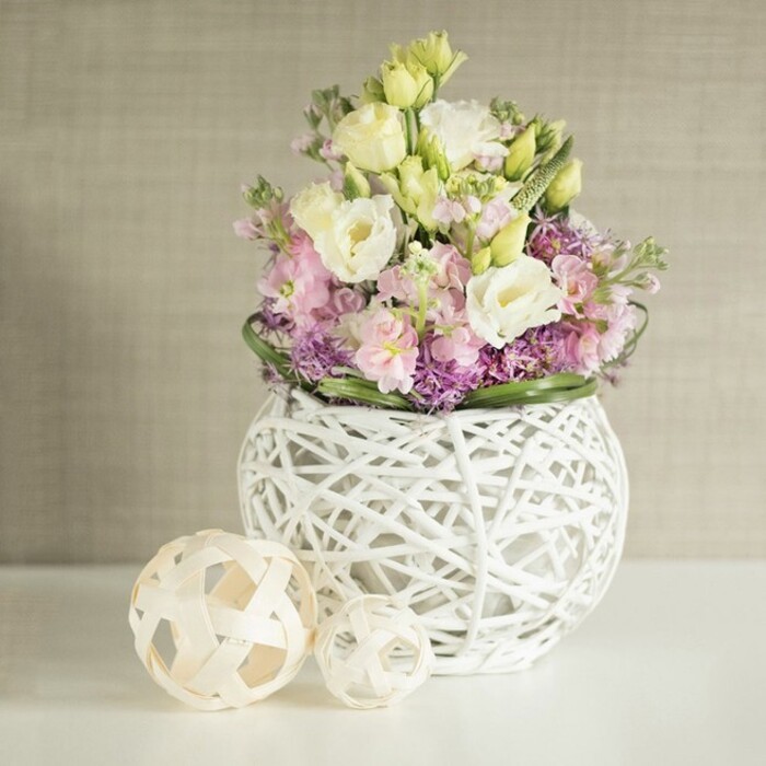 A graceful, painterly scene unfolds on a white table inspired by Clerkenwell's eclectic elegance, as an abundant arrangement emerges in soft, inviting tones. Dominating the composition, creamy white and pale yellow eustoma blossoms-lisianthus-unfurl their layered, ruffled petals in a radiant display, while vertical accents from pink and lavender matthiola stocks draw the gaze upward, imparting buoyancy and joy. Interwoven below, jewel-toned purple filler flowers provide a plush foundation and depth, reminiscent of the floral splashes in St James's Churchyard. Lush, deep green leaves encircle the perimeter, a natural frame that brings harmony to the range of pastels and purples above. These blooms are artfully cradled in a modern, sphere-shaped vase constructed from an open braid of painted rattan, its airy, geometric weave introducing intrigue and a tactile, contemporary edge. On the table beside the arrangement, twin decorative orbs made from natural, light wood accentuate the scene's organic theme and reflect Clerkenwell's tradition of artisanal craftspeople. The arrangement is set against a calm, vertically textured beige wall, its subtlety allowing the colours to shine without distraction. This tranquil, inviting setting evokes a sense of creative renewal and bespoke beauty-perfect for inspiring events, wedding centrepieces, or thoughtful gifts for homes and studios throughout Clerkenwell, all lovingly prepared by your local florist team.
