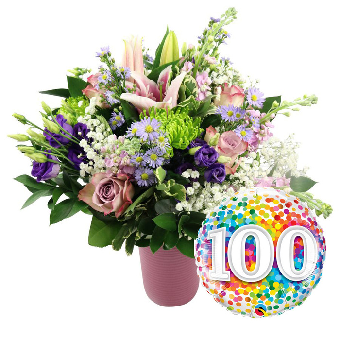 A radiant 100th birthday bouquet and balloon stand against a clean white background, the kind of celebratory display you might find ready for delivery from Clerkenwell Flowers to a townhouse near Clerkenwell Green. The arrangement is gathered into a contemporary ribbed vase in soft matte pink, its cylindrical shape and subtle texture giving a modern, understated base for the exuberant flowers above. At the centre of the bouquet, light pink Oriental lilies take pride of place, their wide, star-shaped blooms facing outward, while several buds reach upwards, still wrapped in pale green and blush, suggesting new chapters in a long life. Surrounding them, soft pink roses and deeper dusty rose blooms unfurl layer by layer, adding romance and a sense of time-honoured elegance. Darker purple lisianthus flowers tuck themselves between the roses, their silky petals creating pockets of rich colour that help the eye travel around the design. Bright lime green button chrysanthemums offer playful, rounded bursts of texture, contrasting with the dainty, feathery clusters of white baby's breath that drift like light clouds between the stems. Sprays of lavender asters, each with a vivid yellow centre, introduce a gently wild, garden-inspired note, as if the bouquet has captured a piece of a late-summer border. Glossy, deep green foliage supports the blooms, filling in gaps and giving the overall shape a lush, abundant feel. To the lower right, partially overlapping the pink vase, a round foil balloon gleams. Its surface is densely patterned with multi-coloured confetti spots in red, orange, yellow, green, blue, and purple, and at its heart sit large, crisp white numerals reading 100, slightly shadowed for dimension. Together, the bouquet and balloon convey a mood of heartfelt celebration, perfect for honouring a centenarian or marking a hundred-year milestone within the close-knit Clerkenwell community.