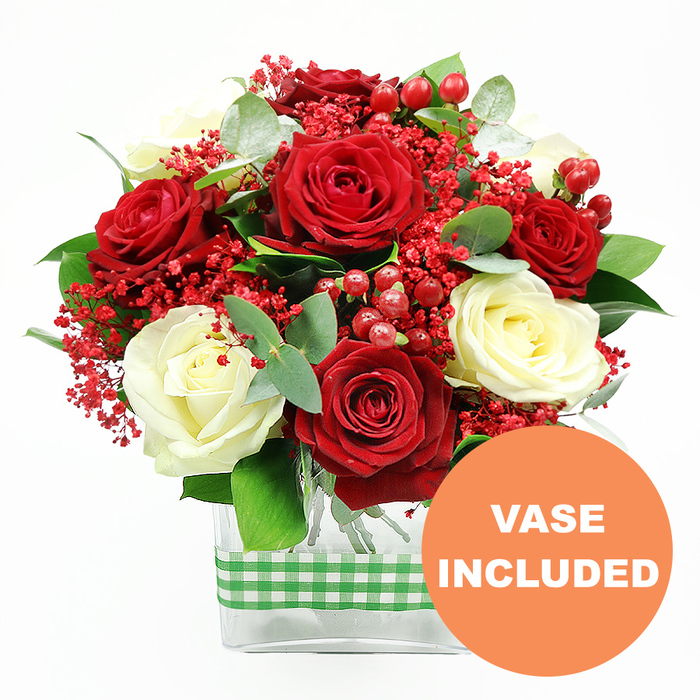 This photograph presents a lush, carefully composed bouquet in classic festive colours, positioned against a crisp white background so every stem and petal is clearly defined. The central focus is a generous grouping of deep red roses, their petals dense and velvety, opened just enough to show intricate spirals. Intertwined with them are creamy white roses, adding softness and balance, their ivory tones glowing gently beside the richer crimson blooms. Filling the spaces between are delicate clusters of tiny bright red filler flowers, similar to dyed gypsophila, which create a fine, airy texture that lightens the arrangement. Rounded, glossy red hypericum berries are tucked throughout, giving the piece a cheerful, berry-laden look that feels particularly suited to winter celebrations or intimate dinners in Clerkenwell townhouses. Surrounding and supporting the blooms, mixed greenery and sprigs of eucalyptus provide structure and depth, the cool green tones and rounded leaves introducing a fresh, natural contrast. All of the stems are gathered into a modern, clear rectangular glass vase, with the submerged green stems visible through the transparent sides, emphasising the clean lines and contemporary style. Around the middle of the vase, a band of green and white gingham ribbon encircles the glass, adding a charming country note that contrasts gently with the urban character of nearby Farringdon and Old Street. In the bottom right corner of the image, an orange circular graphic stands out against the white background, marked with crisp white letters spelling VASE INCLUDED, signaling that the bouquet is delivered exactly as shown, ready to enjoy immediately as a romantic gift, a seasonal centrepiece, or a simple expression of affection delivered across Clerkenwell.