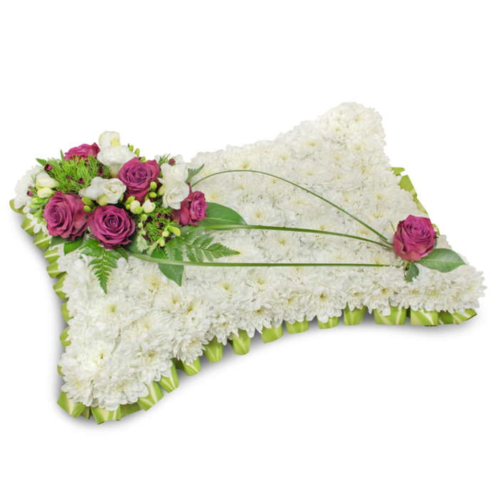 A traditional pillow-shaped bereavement tribute, meticulously assembled from a dense mattress of immaculate white chrysanthemum florets that create a seamless, cloudlike surface against a pure white studio background and faint, grounding shadow. The upper-left is graced by a composed bouquet of deep magenta roses-each bloom plush and velvety-intermingled with petite white freesia or creamy lisianthus that lend a gentle perfume and lightness to the grouping. Around the focal cluster, abundant green foliage-feathery fern fronds and glossy leaves-fans out to enhance depth and to set the roses in a natural frame. Across the white chrysanthemum field, three slender dark-green bear grass stems arc with poised movement, elegantly connecting the dominant spray with a solitary magenta rose nestled near the lower-right corner, a thoughtful counterpoint that completes the visual dialogue. The pillow's edge is wrapped in a neat pleated ribbon of light lime satin, forming a delicate scalloped border that softens the silhouette and reinforces the cushion metaphor. The overall palette-pure white, jewel-toned magenta, and fresh green-reads as dignified and consoling. Picture this piece placed at a memorial in Farringdon or left beside a quiet chapel bench in Spa Fields; its textures invite touch while its restrained scent of rose and freesia offers a discreet, comforting presence. A floral tribute such as this speaks of final rest and respectful remembrance, made with the steady hand of a local craftsman who knows the pace and privacy of Clerkenwell's services.