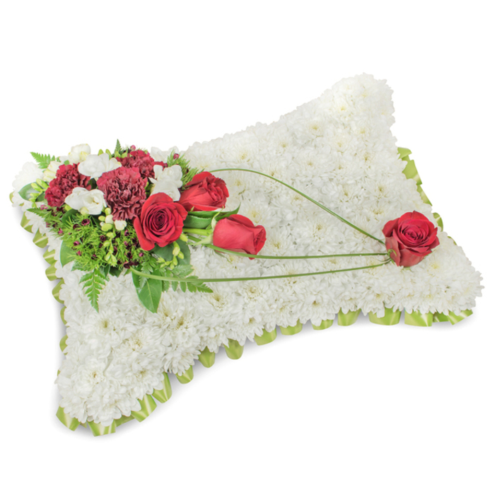 A poised and reverent floral cushion is depicted in soft, even light, its rectangular silhouette formed from hundreds of compact white chrysanthemum heads that together create a dense, velveteen surface reminiscent of a soft shroud. The chrysanthemum field reads as luminous white, offering an impression of calm and purity, while a subtle rim of light lime-green ribbon or foliage traces the pillow's contour, giving definition and a hint of spring-like freshness. Anchoring the composition at the upper left is an opulent spray of red-roses in various stages of bloom, their velvety petals deep crimson, partnered with ruffled burgundy carnations and clusters of small white accent blooms such as freesias that add delicate points of contrast. Rich green foliage, including airy fern fronds and sturdier leaves, fans out behind the floral cluster to provide depth and a living backdrop. From this corner the florist has woven three long, sinuous blades of bright green grass or decorative ribbon across the white surface, their elegant curves drawing attention toward the lower right where a single flawless red rose rests as a quiet focal point-an emblem of lasting affection. The arrangement's balance of texture and colour, its measured diagonal movement, and the restrained yet expressive use of red on white create a soothing presence suitable for funerals, memorial services, or placing at a graveside. One can imagine it arriving with gentle handling to a funeral home near Mount Pleasant or left beside a chapel table in Spa Fields, its craftsmanship speaking to deep care and thoughtful remembrance.