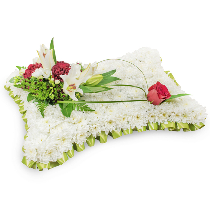 A timeless sympathy cushion shaped into a neat rectangle, completely covered with hundreds of small white chrysanthemums whose tightly layered petals produce a soft, even surface reminiscent of a restful pillow. The perimeter is finished with a pale lime green satin ribbon, carefully pleated to form a delicate, fabric-like frill that softens the silhouette and introduces a gentle hint of colour. On the upper left quadrant a cluster of floral accents rises with quiet dignity: two elegant white Oriental lilies open to reveal pale yellow stamens, balanced by a single closed lily bud and several deep burgundy spray carnations that lend warmth and depth. The arrangement is supported by a variety of green foliage - fern-like textures and broader glossy leaves - which create a layered, natural backdrop. Two slender, sweeping strands of green foliage or decorative wire extend from the cluster across the cushion, guiding the eye to a solitary deep red rosebud poised on the upper right edge, its petals beginning to part as if to speak of memory and enduring affection. The lilies give a soft, floral scent that mingles with the cool, clean presence of the chrysanthemums, producing a composed, reverent aroma. Photographed on a white field with soft, even daylight, this floral pillow is a dignified expression of condolence, suitable for memorial services in local venues around Clerkenwell Green, Mount Pleasant, or the nearby parish, conveying solace and respect to mourners.