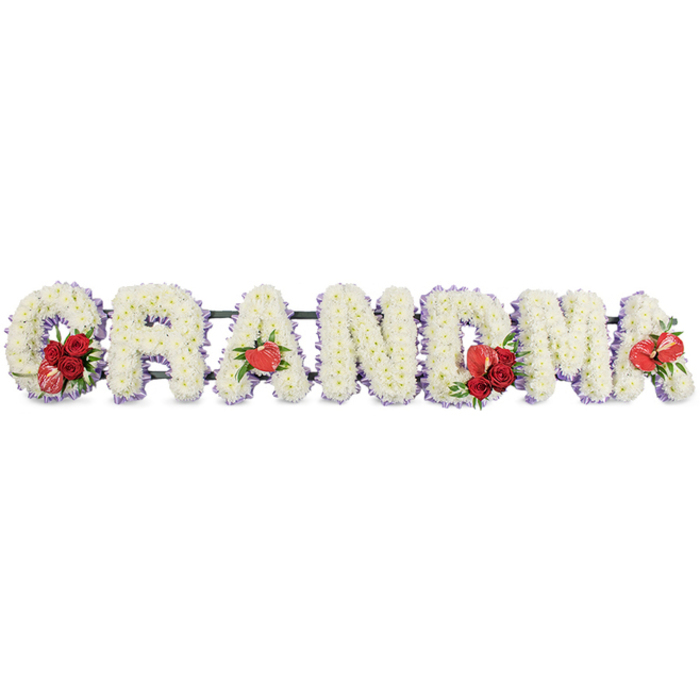 A large funeral flower tribute spells out the word GRANDMA in individual block letters, each one lovingly constructed by hand and displayed against a clean, uncluttered white background. The letters are generously filled with white cushion chrysanthemums, or similar dainty blooms, packed so densely that not a gap of the base shows through, giving the surface a plush, quilted look. Around the outer edge of each capital, a trim of soft lilac or light purple ribbon peeks through, framing the flowers and adding a gentle suggestion of colour that feels calm and comforting. Rich red floral accents are arranged with intention rather than symmetry: at the front of the G, a spray of velvety red roses and a distinctive red-and-cream variegated calla lily nestle among glossy greenery, like a small hand-tied posy set into the shape. The first A is anchored with one shining red anthurium at its foot, its heart-like form drawing the eye. In the middle of the tribute, the M bears another grouping of red roses paired with a second red anthurium, and the final A completes this rhythm with a single red anthurium bloom. Beneath the letters, a dark green structural frame provides support, allowing the tribute to stand or be propped up on an easel during a service in Clerkenwell, perhaps near St John Street or close to local crematoria used by City families. The overall impression is one of quiet strength and tenderness, a bespoke floral message saying GRANDMA in a way that feels both traditional and deeply personal, suitable for honouring her memory at a funeral, wake, or graveside gathering.