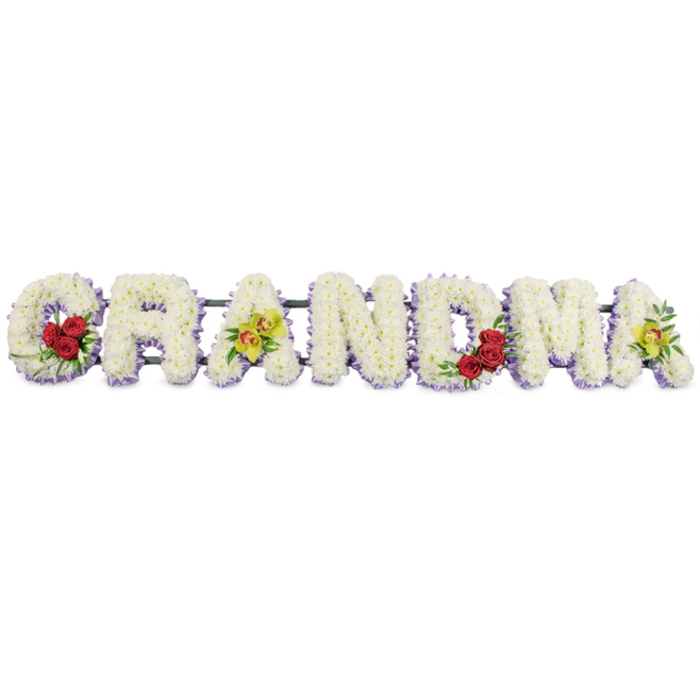 In this detailed sympathy design, the word GRANDMA is formed from separate floral letters arranged neatly in a row, photographed head-on against an uncluttered white background that emphasises their sculpted shapes. Each capital letter is built on a sturdy frame and completely covered with compact white chrysanthemums, the blooms nestled so closely that no gaps of greenery are visible, giving the letters a cushiony, uniform texture. Just along the lower edges, a subtle band of soft lavender flowers peeks through, lightly brushing each letter with a cool hint of colour and enhancing the definition of the word. At the left side, the G is distinguished by three rich red roses gathered on its lower curve, their velvety petals unfurling in deep tones of crimson and framed by polished green leaves. The first A is embellished with two lime-green cymbidium orchids poised on the crossbar, their intricate centres flecked with warm reddish markings that draw the eye. The middle letters R, N, and D are left purely white with the lavender trim, creating a calm, steady rhythm across the tribute. Closer to the right, the M cradles another small group of three red roses in its lower right dip, thoughtfully mirroring the G to provide visual balance. The final A repeats the signature yellow-green orchids, echoing the first and gently closing the design. A slim, dark metal bar runs behind the top of the letters, mostly hidden, providing structure so the piece can stand as a focal point at a farewell gathering in Clerkenwell, perhaps in a quiet chapel or civic space near Clerkenwell Green. The piece evokes tenderness and respect, a personalised farewell for a beloved grandmother whose presence shaped generations.
