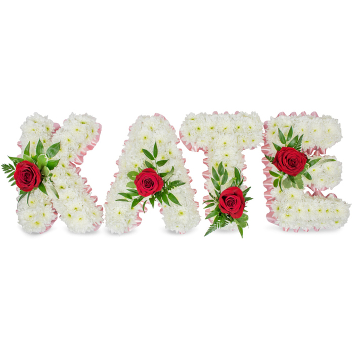 A meticulously crafted memorial piece presents the name KATE in voluminous block letters, each letter constructed from a thick, uniform layer of small white chrysanthemums whose densely packed petals create an even, cloud-like texture. The edges of every letter are trimmed with a fine, pale pink pleated satin ribbon that follows the letterforms precisely, lending a soft framing that warms the stark white without overpowering it. A single, richly coloured red rose punctuates the face of each letter as a central accent - positioned on the lower left vertical of the K, at the top peak of the A, at the left base of the T's crossbar, and on the middle arm of the E - each rose nestled amid a generous sweep of dark green foliage, from broad glossy leaves to delicate fern fronds that add movement and depth. Against a clean white backdrop the composition reads with calm clarity, a traditional sympathy tribute that balances formal structure with tender detail. The roses' velvety petals and the chrysanthemums' subtle texture are highlighted by soft directional lighting, and there is a faint, comforting scent that seems to belong to quiet rooms and remembrance. As a piece one might commission for a funeral in Clerkenwell or a nearby chapel off Leather Lane, the arrangement demonstrates the measured craft of a local florist: considered proportions, clean finishing, and an intent to honour with dignity and warmth.