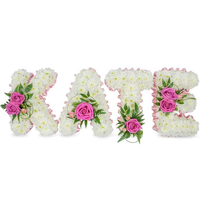 This bespoke memorial piece spells the name KATE in generous three-dimensional letters, each letter constructed from hundreds of small, immaculate white chrysanthemums tightly clustered to create a soft, voluminous texture that reads like a cloud. A narrow pale-pink satin ribbon delicately outlines the inner contours of every letter, giving a refined, feminine accent to the sculpted florals. Carefully placed clusters of hot-pink roses bring visual punctuation: the K is adorned with two open roses on its left side framed by fresh green leaves, the A carries a single prominent rose tucked into its lower curve highlighted by slender green sprigs, the T is finished with one vivid pink bloom at its base among fern-like foliage, and the E displays two roses on its right edge set against variegated leaves and tiny pink floral accents. Under soft studio light the chrysanthemums appear luminous while the pinks read as warm and consoling; the arrangement casts gentle shadows on the white background that emphasise its freestanding, ceremonial presence. Small light-pink buds and delicate spray flowers intersperse the composition, introducing subtle texture and the suggestion of a delicate perfume from the roses. This floral lettering is designed to communicate remembrance, love and respect - a dignified, personalised sympathy display often chosen for memorial services and celebrations of life in Clerkenwell, from St John's Square to the burial chapels around Hatton Garden and Smithfield. The craftsmanship suggests careful work in a neighbourhood workshop, suitable for placement beside a casket or on a commemorative table.