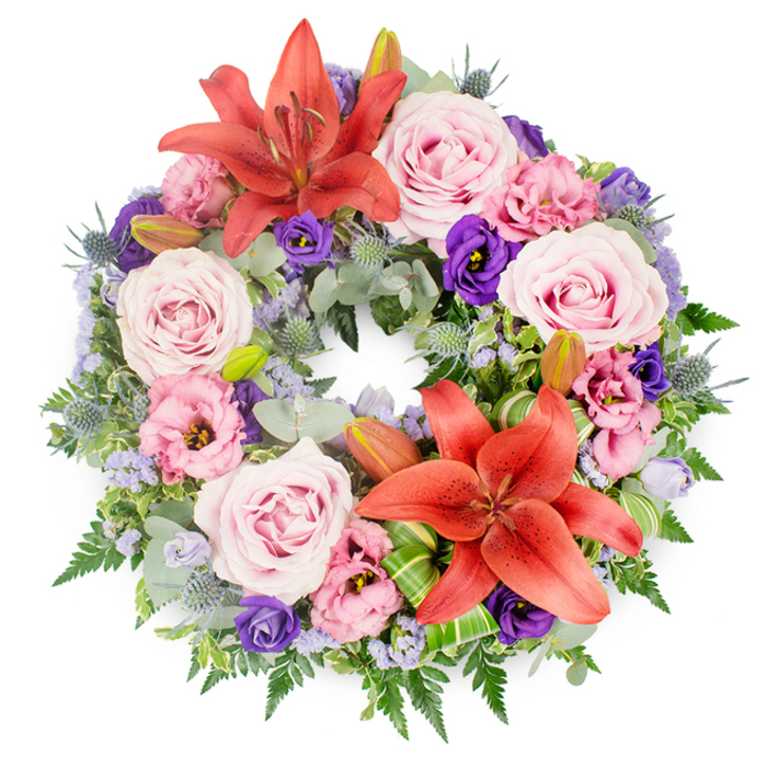 Captured from directly above against a bright white backdrop, this full funeral wreath reveals a meticulously arranged circle of seasonal flowers and foliage. The foundation is an abundant mix of greens: cool, rounded eucalyptus leaves, slim fern fronds, and darker, glossy foliage that together create a deep, living cushion. Resting upon this are generous clusters of roses in soft blush and lavender-pink, their petals spiralling open to show every velvety layer. Interspersed with them are ruffled lisianthus blooms, some in a powdery pale pink and others in a saturated purple tone, which lend the tribute both delicacy and depth. Two large Asiatic lilies in a striking coral-orange hue serve as focal points at opposite points of the circle, their star-shaped flowers fully open, with stamens clearly visible and a faint suggestion of fragrance drifting from their centres. Around them, a few unopened lily buds extend upwards, symbolising life continuing beyond the moment of farewell. Spiky blue-grey eryngium thistles punctuate the arrangement with a sculptural, almost metallic texture, while wisps of lavender-blue statice thread through the design like a gentle, airy veil. Near the lower lily, folded and pleated bright green leaves, likely aspidistra or ti leaves, demonstrate expert floristry and add a modern, structured touch. The overall palette of pinks, purples, oranges, greens, and soft blues feels harmonious rather than bright, making the wreath an elegant choice for a memorial or funeral service in Clerkenwell. Whether placed by a chapel door off St John Street or travelling with a hearse through the local streets, it conveys peace, gratitude, and enduring affection.