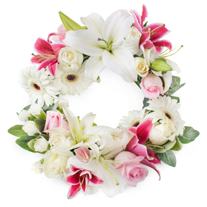 This photograph shows a full, circular funeral wreath laid out against a pristine white background, the design rich with white, blush, and deep magenta flowers arranged in a seamless ring. Bold white lilies form the foundation, their broad petals opening outward with a quiet grace, each flower revealing central stamens brushed with warm gold and soft green pistils that lend a sense of natural calm. Among them, deep pink and fuchsia lilies add depth and emotion, their velvety petals gently speckled and slightly curled at the tips, suggesting both beauty and farewell. White gerbera daisies are scattered throughout the circle, their neat, layered petals surrounding dark centres that provide striking contrast, ideal for a modern memorial in Clerkenwell's chapels or civic spaces. Soft pink roses appear in varying stages-some tight buds, others just beginning to unfurl-while cream and ivory roses open fully, their layered petals creating a sense of abundance and care. Fine white filler flowers, possibly miniature carnations or sprays of chrysanthemum, are tucked between larger blooms so that every section of the wreath feels intentional and complete. A framework of deep green foliage supports the entire design, leaves peeking through to add texture and a natural outline to the ring. The empty centre of the wreath offers a quiet focal space, symbolising remembrance and the continuation of life's circle. The overall feel is one of dignified tenderness, a tribute that could comfortably rest in a church near Clerkenwell Green, a crematorium chapel, or a family home, bringing comfort and a touch of floral beauty during a time of loss.