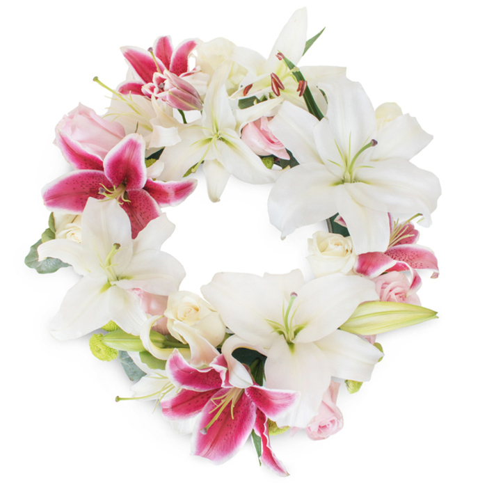 Presented against a crisp white backdrop, this opulent floral wreath exemplifies the artistry found at Clerkenwell's local florist studios. The design features a lush circle densely adorned with large, pristine white Oriental lilies, whose broad, slightly curved petals open gracefully to reveal subtle green pistils and gentle yellow stamens. Contrasting these luminous whites are several fuchsia-pink stargazer lilies, their vivid, saturated petals peppered with darker speckles and edged in soft blush, imparting a dynamic visual rhythm and infusing warmth into the scene. Cozy between the statement lilies, small clusters of creamy blush and pale pink roses-some tightly furled, some just opening-provide a delicate texture reminiscent of rose beds in local gardens along Rosebery Avenue. Discreet hints of muted green foliage, evocative of eucalyptus or fresh-cut botanical greens from Exmouth Market, lend structural form, while subtle shadows create depth and the illusion of freshly gathered blooms in morning light. The full, continuous ring and the open central space of the wreath speak to eternal remembrance and communal unity, inviting a moment of reflection. Every detail is thoughtfully balanced, conveying sympathy and dignity; a perfect centrepiece for Clerkenwell memorials, farewells, or as a bespoke condolence tribute, expertly arranged by seasoned hands deeply rooted in the neighbourhood.