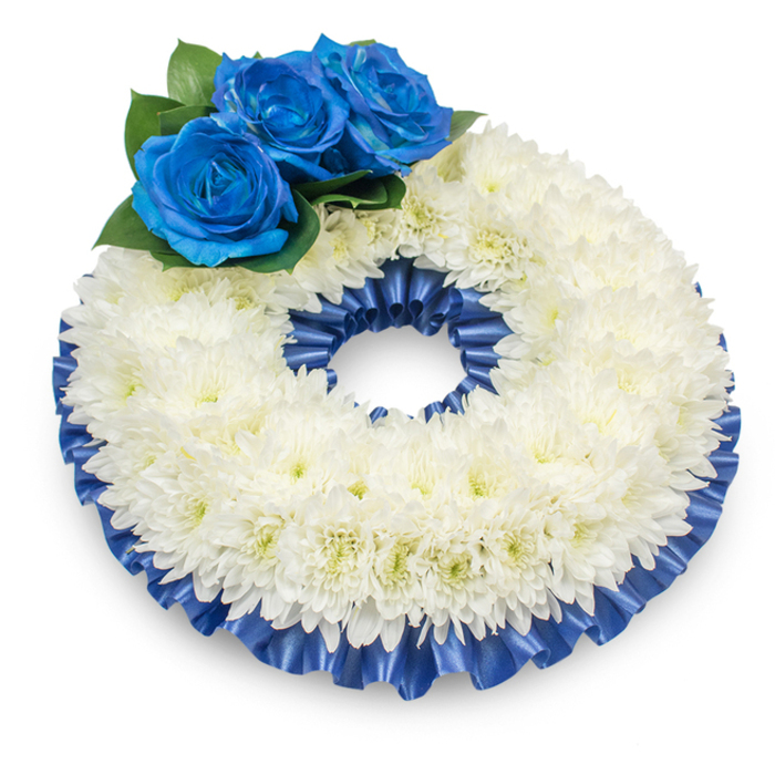 This circular wreath is portrayed against a bright white background from a modestly elevated angle so every element reads clearly: a dense, continuous ring of pristine white chrysanthemums creates a soft, cloud-like mass with palpable texture, the thousands of feathery petals layered to form a slightly domed surface that speaks of careful, patient handwork. The chrysanthemum centres show a subtle creamy tint that warms the composition while the main impression remains peaceful and pure. Placed atop the upper left section are three vivid, deep electric-blue roses, their petals richly saturated and velvety to the eye, arranged among rounded, dark green foliage which accentuates the blue and helps the cluster gently emerge from the white background. A pleated cobalt satin ribbon trims both the inner and outer edges of the chrysanthemum ring; its ruffled folds and glossy finish give a refined frame, catching light along its pleats to create soft highlights that enhance the wreath's formal silhouette. The lighting produces a delicate shadow beneath the arrangement, lending it weight and a sense of presence as if ready for a graveside or chapel setting. The tactile contrasts are clear in mind: the cushiony softness of chrysanthemum petals against the smooth sheen of ribbon and the resilient firmness of rose petals. The scent is understated - a clean, comforting chrysanthemum note with a green whisper from the foliage. This composition reads as a respectful floral tribute, appropriate for funerals or memorial services in Clerkenwell and nearby Farringdon, offering comfort through timeless symbolism and considered craftsmanship.