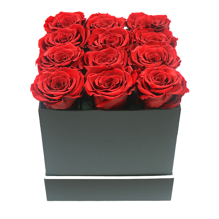 A refined, studio-style photograph showcases a contemporary rose box arrangement that radiates quiet luxury, the sort of piece often requested from Clerkenwell Flowers for significant occasions. Dominating the frame is a compact, square container in a deep matte black, its clean, straight edges giving it a distinctly modern, almost architectural presence. Around the lower third of the box runs a slim, bright white band, like a minimalist belt, adding just enough detail to break the darkness and emphasise the form. Emerging from the open top are twelve lush red roses, precisely arranged in a three-by-four formation so that the blooms touch and interlock, creating a seamless field of velvety petals. Each rose is at its full, open stage, with layered crimson petals unfolding in delicate curls, revealing subtle variations of red-from darker, wine-toned shadows near the centre to slightly brighter edges catching the light. The roses sit slightly above the rim of the box, suggesting fullness and abundance without appearing unruly. They have the immaculate look of preserved or 'eternity' roses, chosen for clients who want a lasting reminder of an anniversary, proposal, or milestone celebration in Clerkenwell. The image is set against a pristine white backdrop that eliminates all context, allowing the arrangement's colour and symmetry to stand out with graphic clarity. Soft, diffused lighting creates gentle highlights along the petal edges and casts faint, controlled shadows beneath the blooms and along the box's sides, adding depth but keeping the mood calm and polished. The overall impression is of a sophisticated, design-conscious gift-ideal for same-day delivery to an office near Farringdon Station, a stylish apartment by Clerkenwell Green, or a discreet romantic surprise sent to one of the area's many creative workspaces.